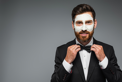 happy and bearded man in suit with clay mask on face adjusting bow tie isolated on grey