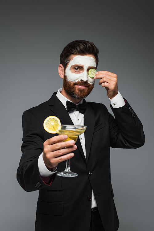 happy man in suit with clay mask on face holding cocktail isolated on grey