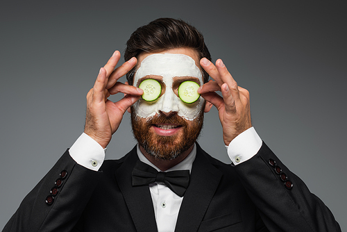 bearded man with clay mask on face applying sliced cucumber on eyes isolated on grey