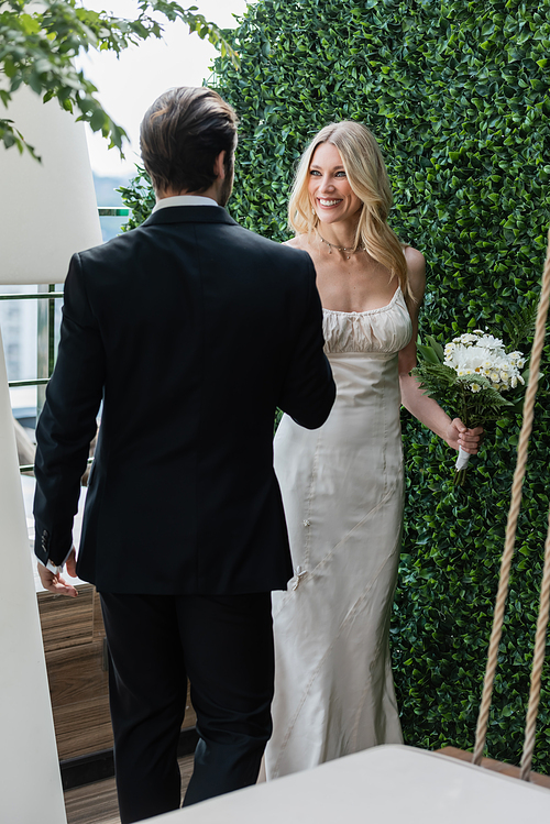 Smiling blonde bride holding bouquet and looking at groom on terrace