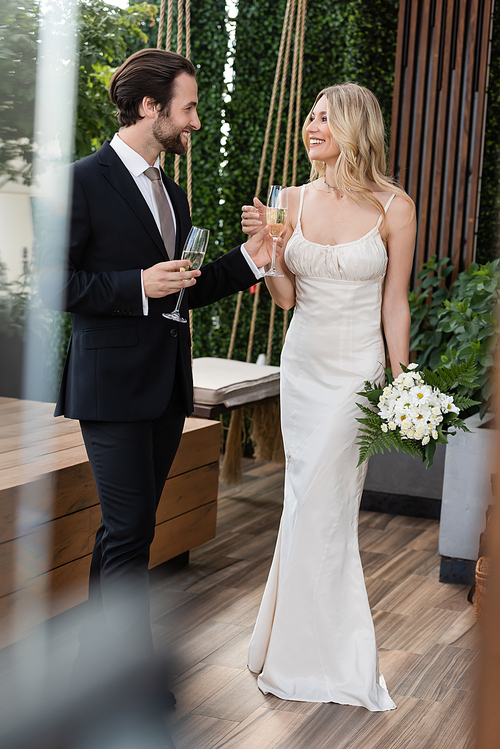 Smiling newlyweds with bouquet and champagne looking at each other on terrace