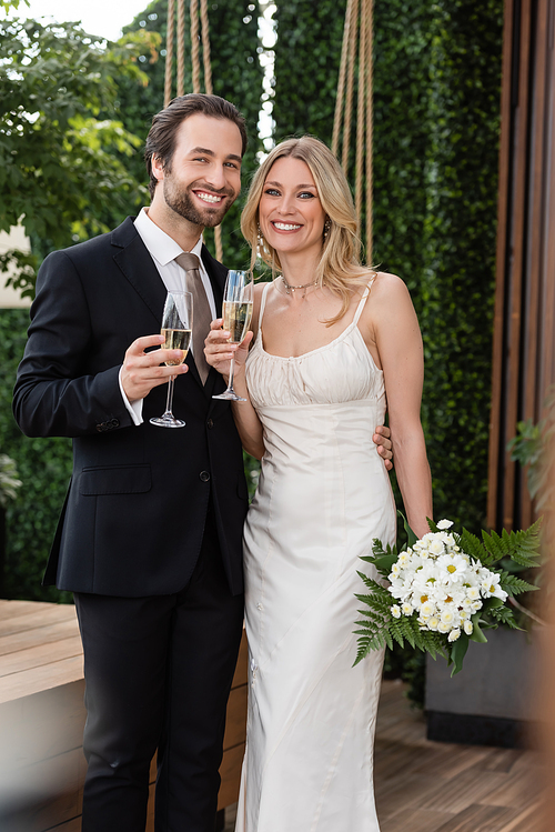 Positive newlyweds with champagne and bouquet looking at camera on terrace