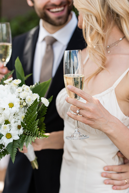 Cropped view of blonde bride holding bouquet and champagne near groom outdoors