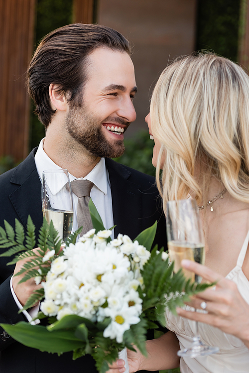 Positive groom holding glass of champagne near bride with bouquet outdoors
