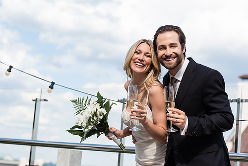Cheerful newlyweds with bouquet and champagne looking at camera on terrace