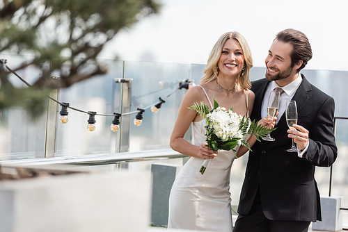 Smiling bride with bouquet and champagne looking at camera near elegant groom on terrace