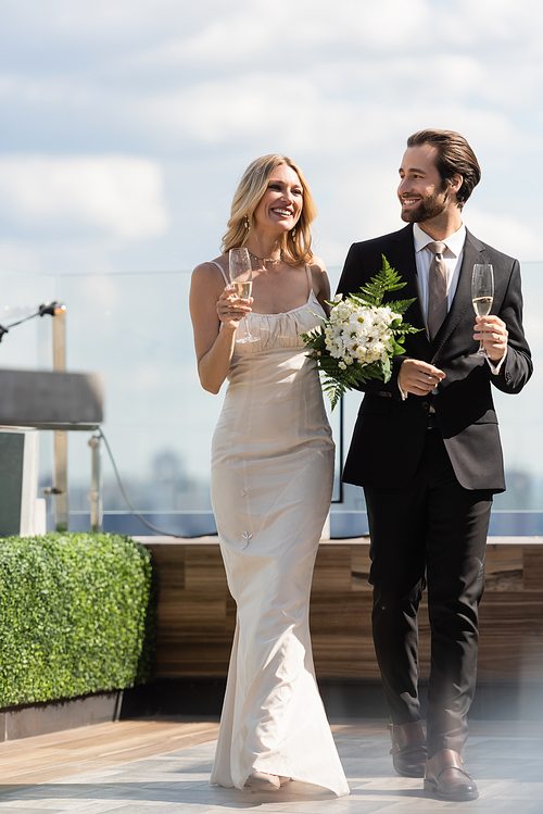 Positive newlyweds with flowers and champagne walking on terrace of restaurant