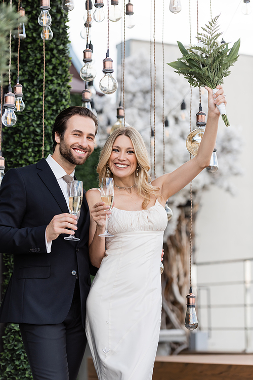 Happy bride holding bouquet and champagne near groom and light bulbs on terrace