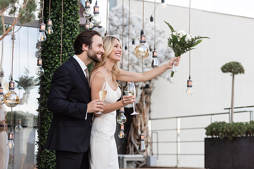 Positive newlyweds with champagne and bouquet standing near light bulbs on terrace of restaurant