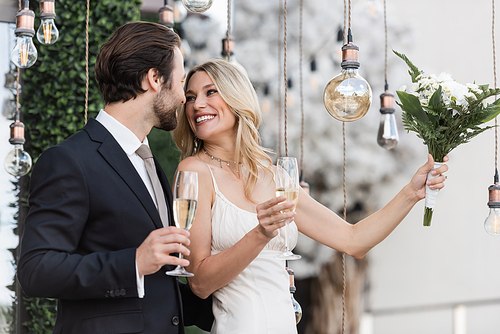 Blonde bride holding bouquet and champagne near elegant groom and light bulbs on terrace