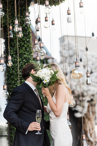 Newlyweds with bouquet and champagne covering faces while kissing under light bulbs outdoors
