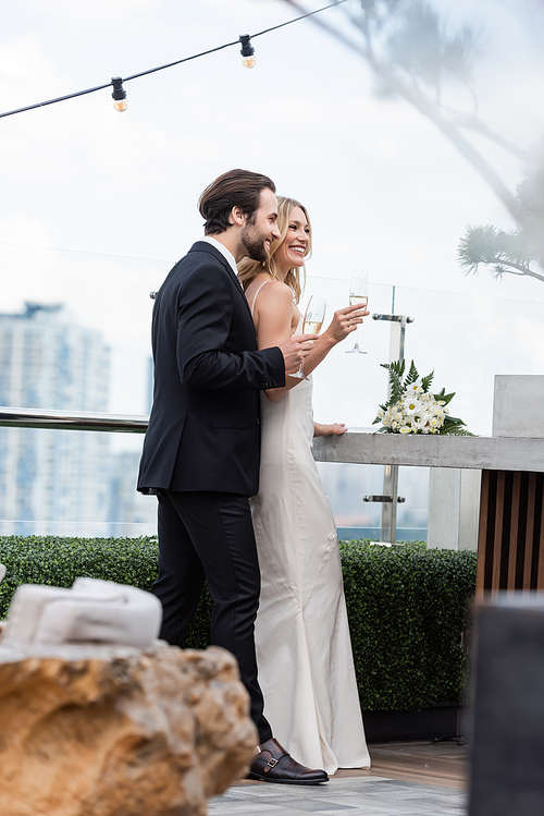 Smiling groom in suit holding glass of champagne near bride and bouquet on terrace