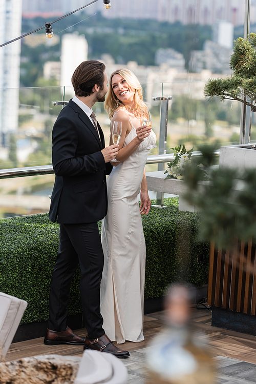 elegant groom holding champagne near smiling bride in dress on terrace