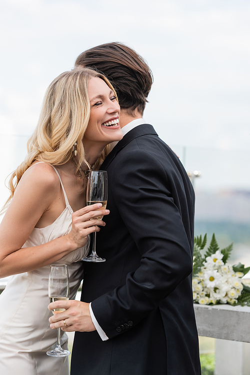 Cheerful bride holding glass of champagne near elegant groom in suit on terrace