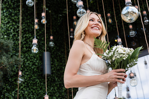 Low angle view of blonde bride holding wedding bouquet near light bulbs on terrace