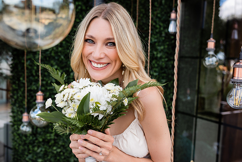 Portrait of positive bride holding bouquet and looking at camera near blurred light bulbs outdoors