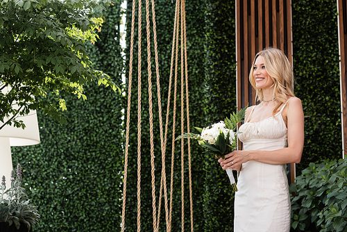 Positive bride holding flowers near plants on terrace