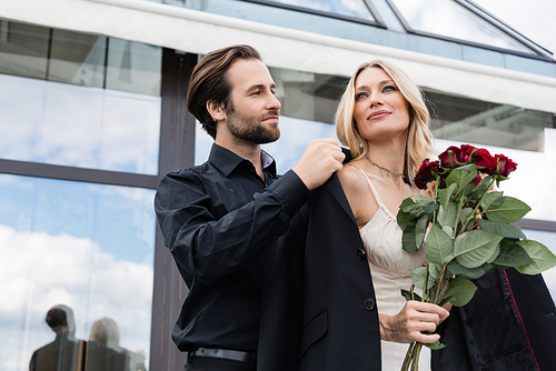 Low angle view of young man wearing jacket on blonde girlfriend with roses during date outdoors