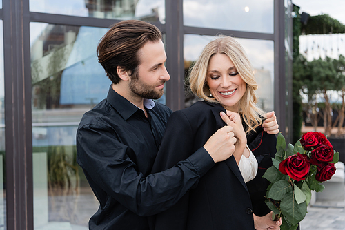 Young man wearing jacket on smiling girlfriend with roses on terrace