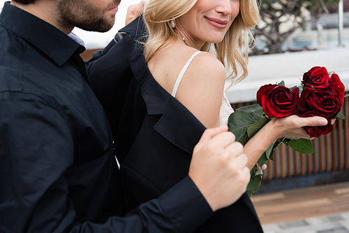 Cropped view of man wearing jacket on blonde girlfriend with roses outdoors
