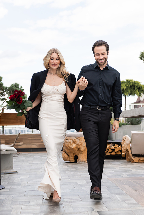 Smiling woman in dress and jacket holding flowers and hand of boyfriend while walking on terrace