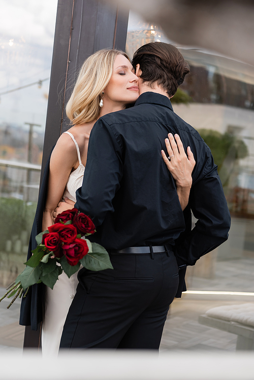 Blonde woman in dress holding red roses and hugging boyfriend outdoors