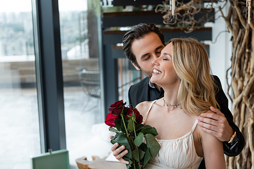 Brunette man kissing cheerful girlfriend with red roses in restaurant