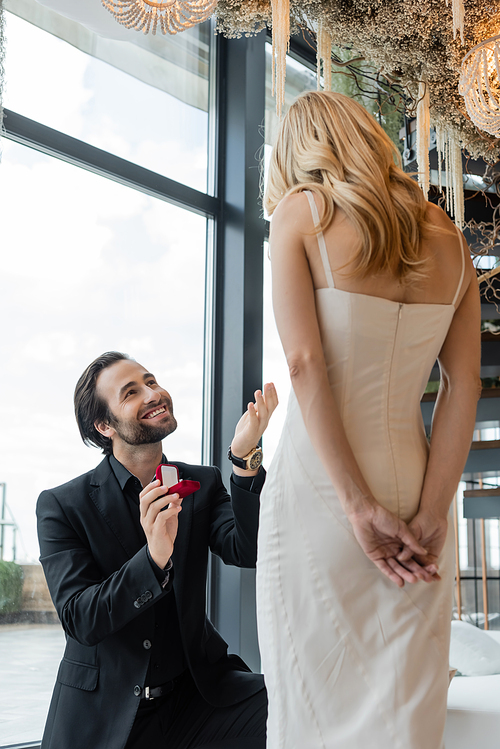 Positive man holding engagement ring near girlfriend in dress in restaurant