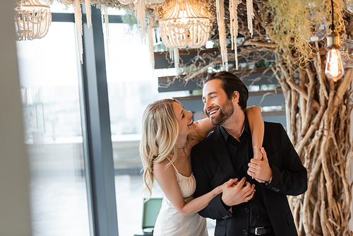 Cheerful woman in dress holding hands of boyfriend under chandelier in restaurant