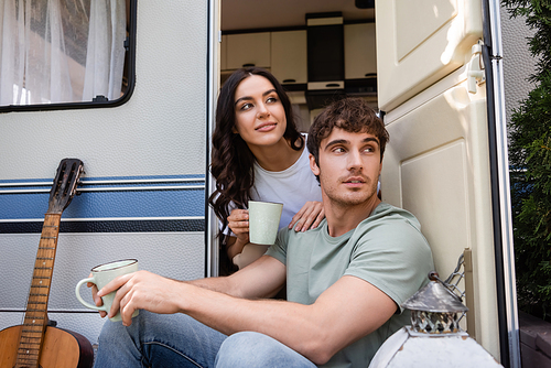 Young couple holding cups of coffee near acoustic guitar and camper van outdoors