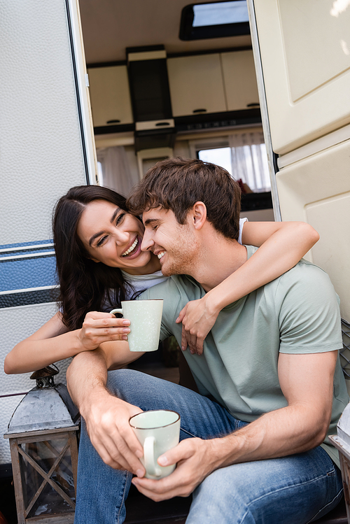 Positive young woman hugging boyfriend with cup near door of camper van