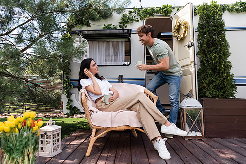 Side view of cheerful man holding cup near girlfriend on armchair and camper van