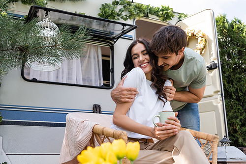 Smiling man hugging cheerful girlfriend with closed eyes holding cup near camper van