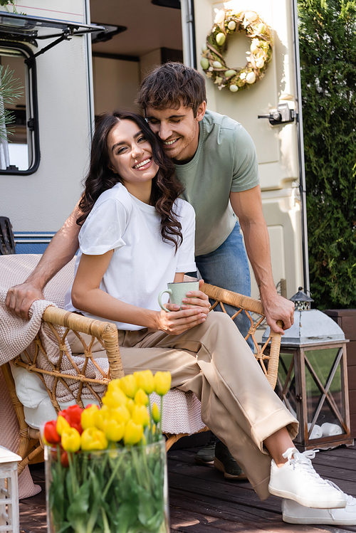 Smiling man standing near cheerful girlfriend with cup on armchair near camper van