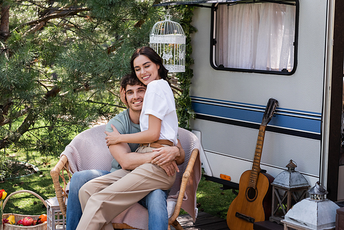 Positive woman hugging young boyfriend while sitting on armchair near camper van