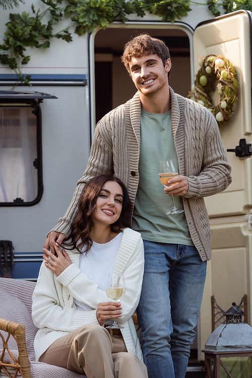 Young couple in warm cardigans holding wine and smiling at camera near camper van