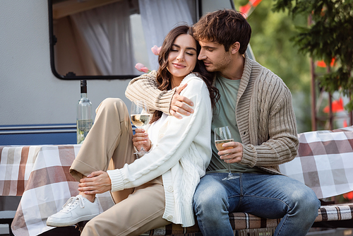 Brunette man in cardigan hugging girlfriend with wine on terrace of camper van