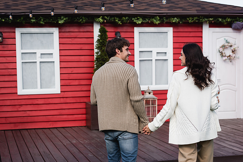 Side view of smiling couple in cardigans holding hands near blurred house