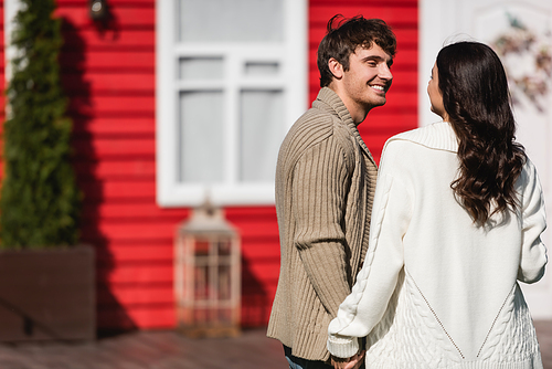 Smiling man in cardigan holding hand of brunette girlfriend near blurred house