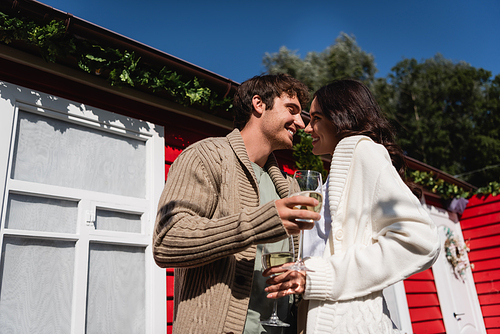 Low angle view of smiling young couple in cardigans holding glasses of wine near house outdoors