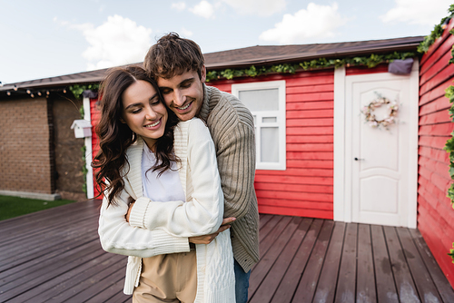 Smiling man in warm cardigan hugging girlfriend near blurred house