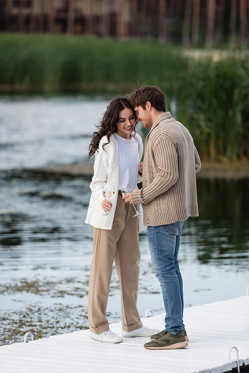 Smiling couple in cardigans holding wine on pier near river