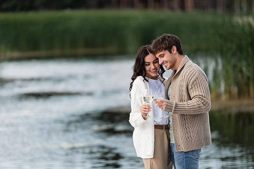 Cheerful couple in knitted cardigans toasting wine near blurred river