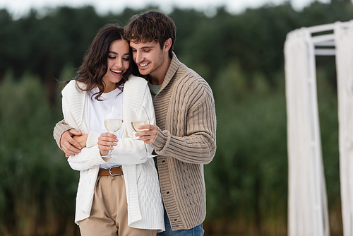 Smiling man in cardigan hugging girlfriend with wine outdoors