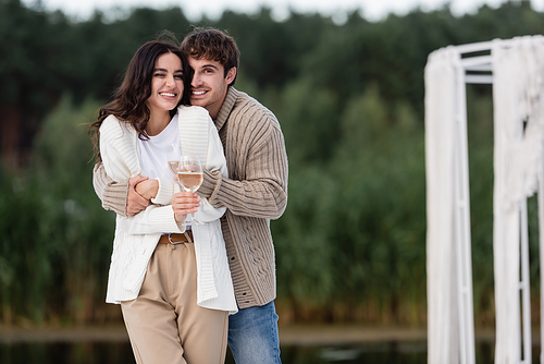Smiling couple in cardigans hugging and holding wine outdoors