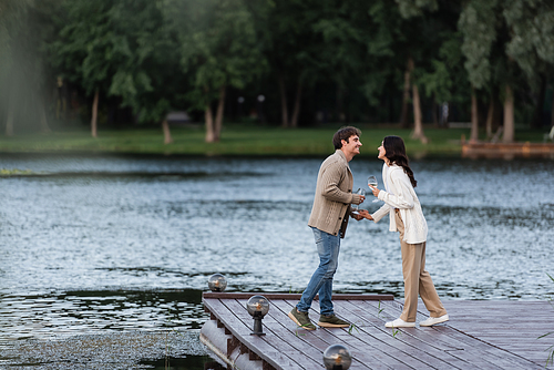 Side view of positive couple in cardigans holding wine on pier near lake