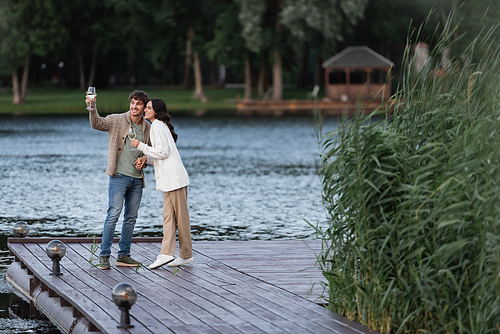 Smiling couple in cardigans holding wine on pier near lake