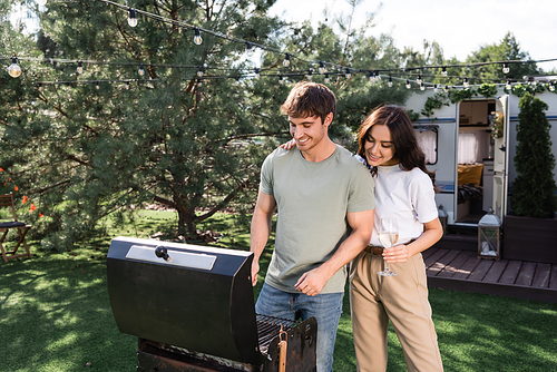 Smiling woman holding wine near boyfriend cooking on grill and camper van