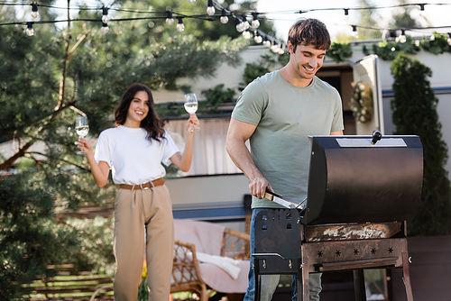Smiling man cooking on grill near blurred girlfriend with wine and camper van