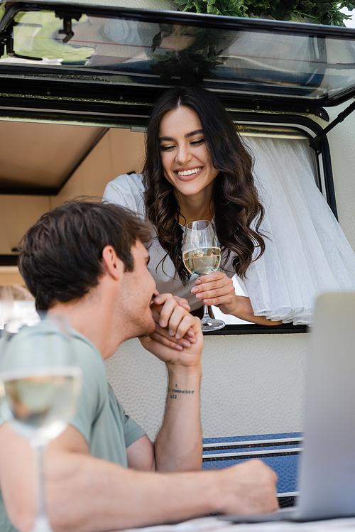 Smiling man kissing hand of girlfriend in camper van near vine and laptop outdoors